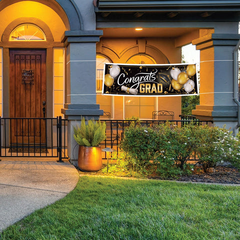Graduation banner with white Congrats lettering gold Grad text and gold silver balloon graphics on black background on a house exterior with decorative elements.